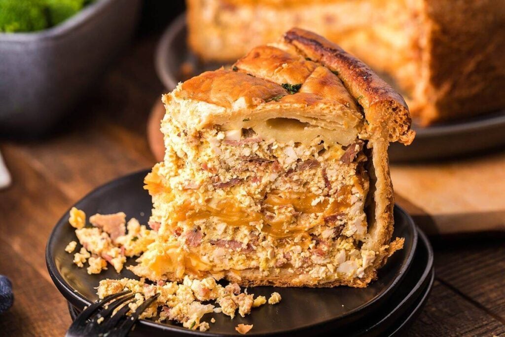 A thick slice of savory pie filled with layers of meat, cheese, and eggs sits on a black plate with a fork; the rest of the pie is visible in the background.