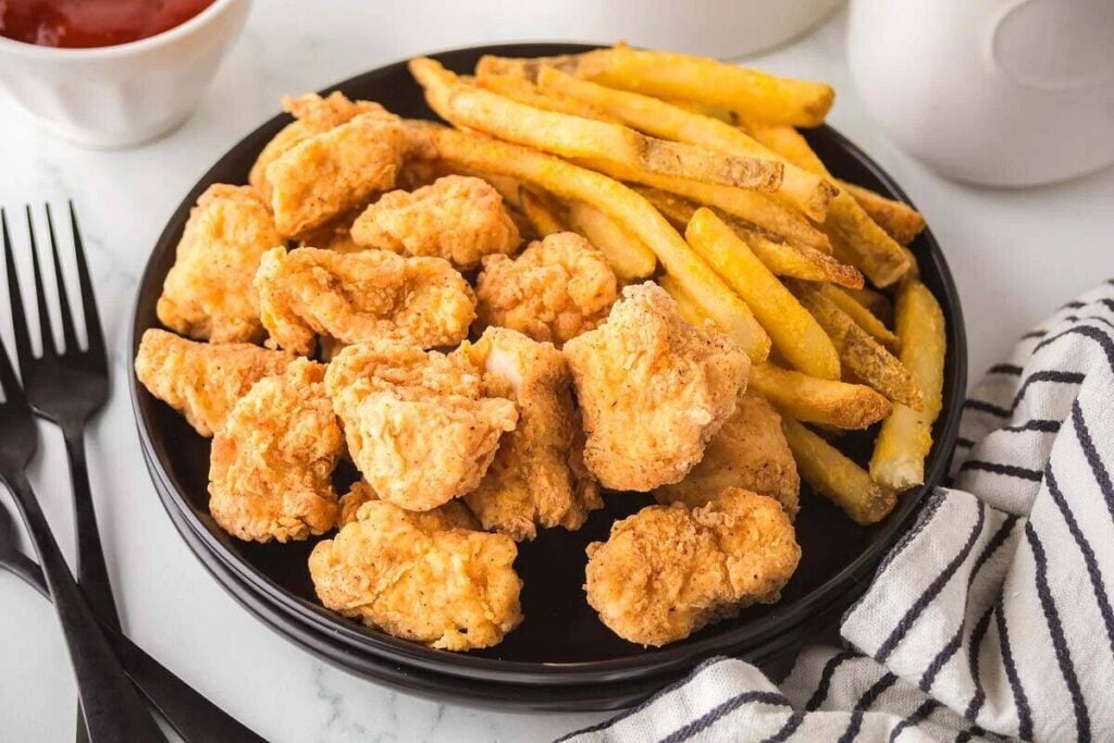 A black plate filled with crispy fried chicken bites and golden fries, next to a striped napkin.