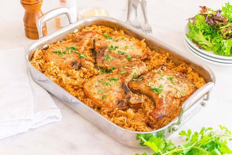 Baked pork chops served on a bed of seasoned rice in a metal roasting pan, with fresh herbs and a side of mixed green salad.