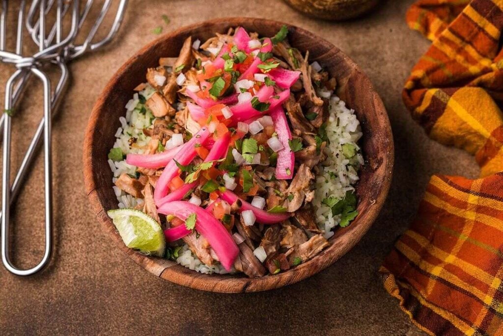 A wooden bowl of rice topped with shredded meat, pickled red onions, chopped cilantro, diced onions, and a lime wedge, set on a brown surface with an orange plaid napkin nearby.