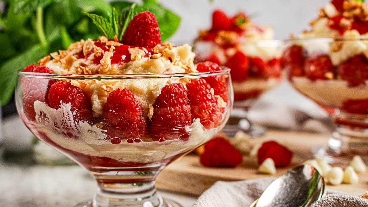 Glass dessert bowls filled with layers of raspberries, cream, and granola, garnished with mint leaves, are arranged on a table with a spoon nearby.