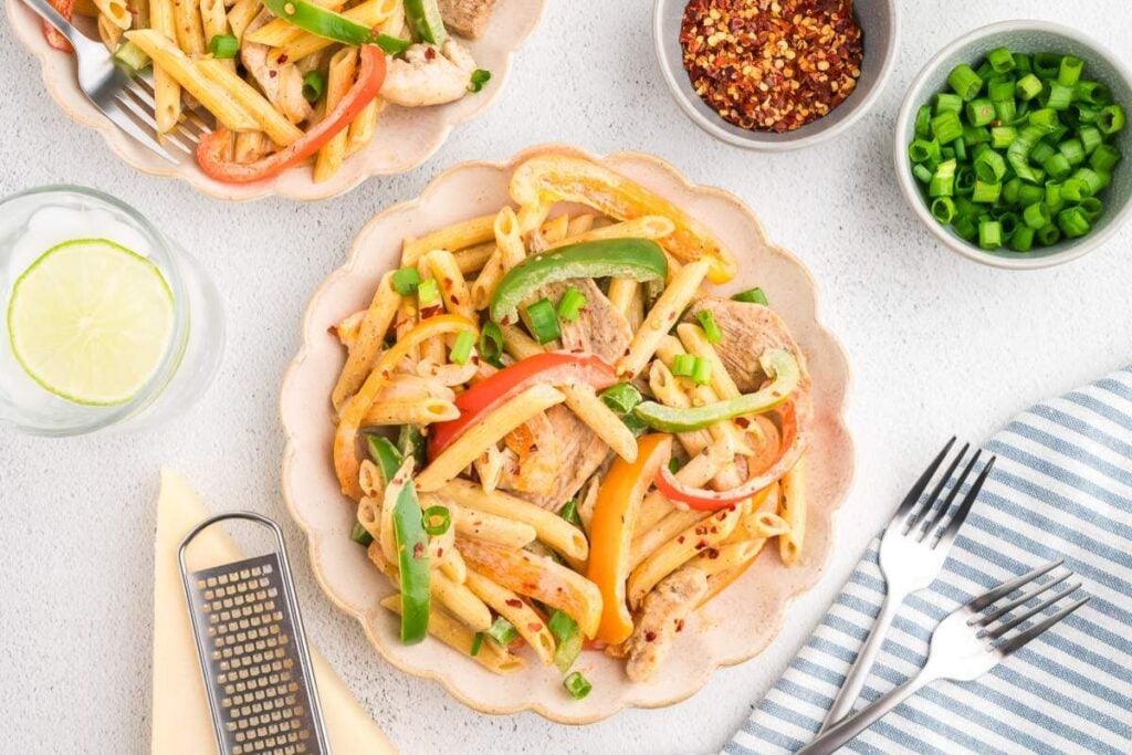 Plate of penne pasta with sliced bell peppers, green onions, and meat, surrounded by bowls of chili flakes and chopped green onions, a glass of water, utensils, and a striped napkin.