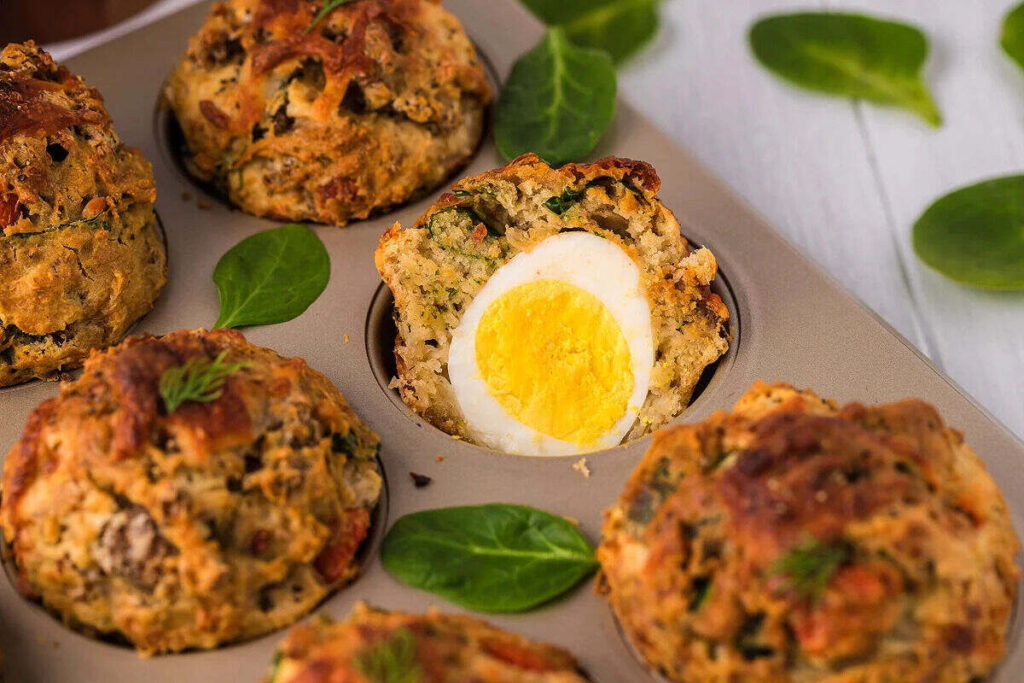 A muffin tin holding savory muffins, with one muffin cut open to reveal a whole boiled egg inside; fresh spinach leaves are scattered around.