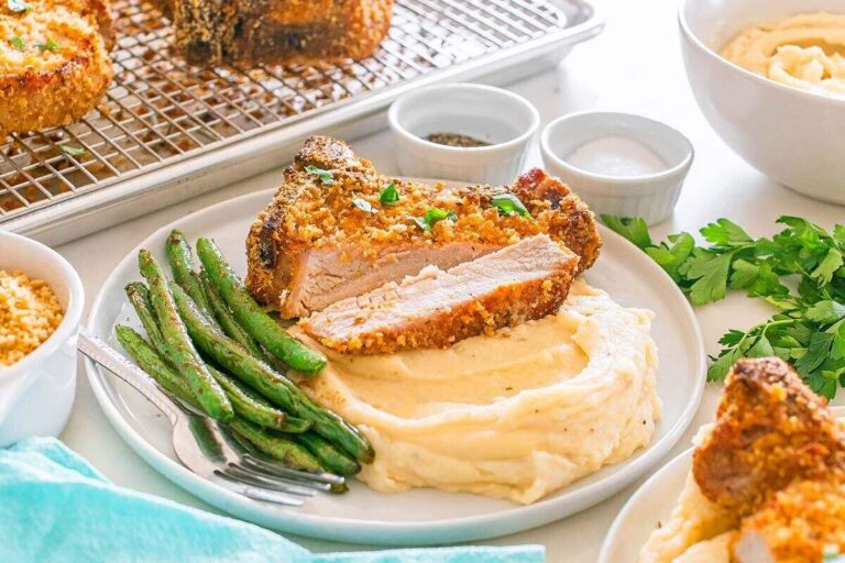 A plate with breaded pork chop slices on mashed potatoes, served with green beans; small bowls of seasonings are in the background.