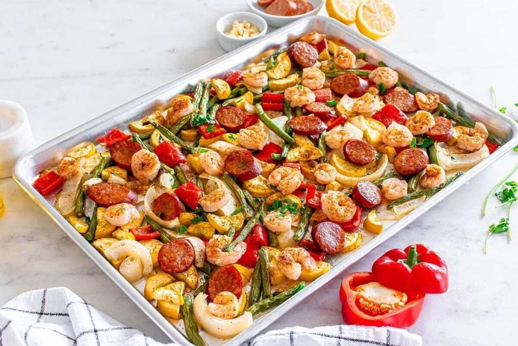 A sheet pan filled with cooked shrimp, sliced sausage, bell peppers, onions, and zucchini, garnished with herbs, sits on a white surface with lemon halves and seasonings nearby.