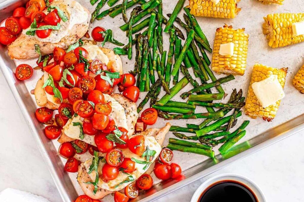 Baked chicken breasts topped with tomatoes and basil, roasted asparagus, and corn on the cob with butter on a sheet pan; cup of dark liquid nearby.