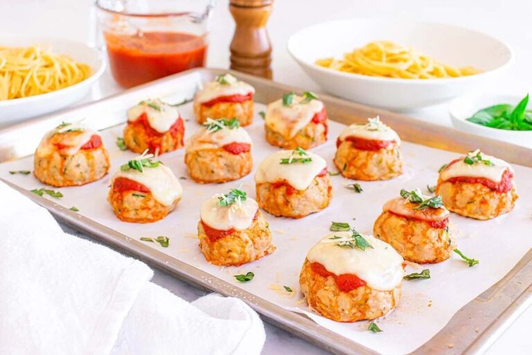A baking sheet with baked meatballs topped with melted cheese and tomato sauce, garnished with herbs. Plates of spaghetti and a jug of sauce are in the background.