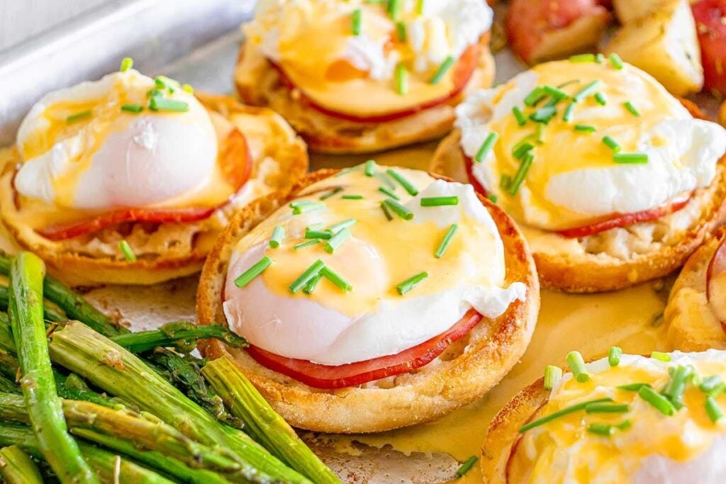 English muffins topped with ham, poached eggs, and hollandaise sauce, garnished with chopped chives, served with roasted asparagus.