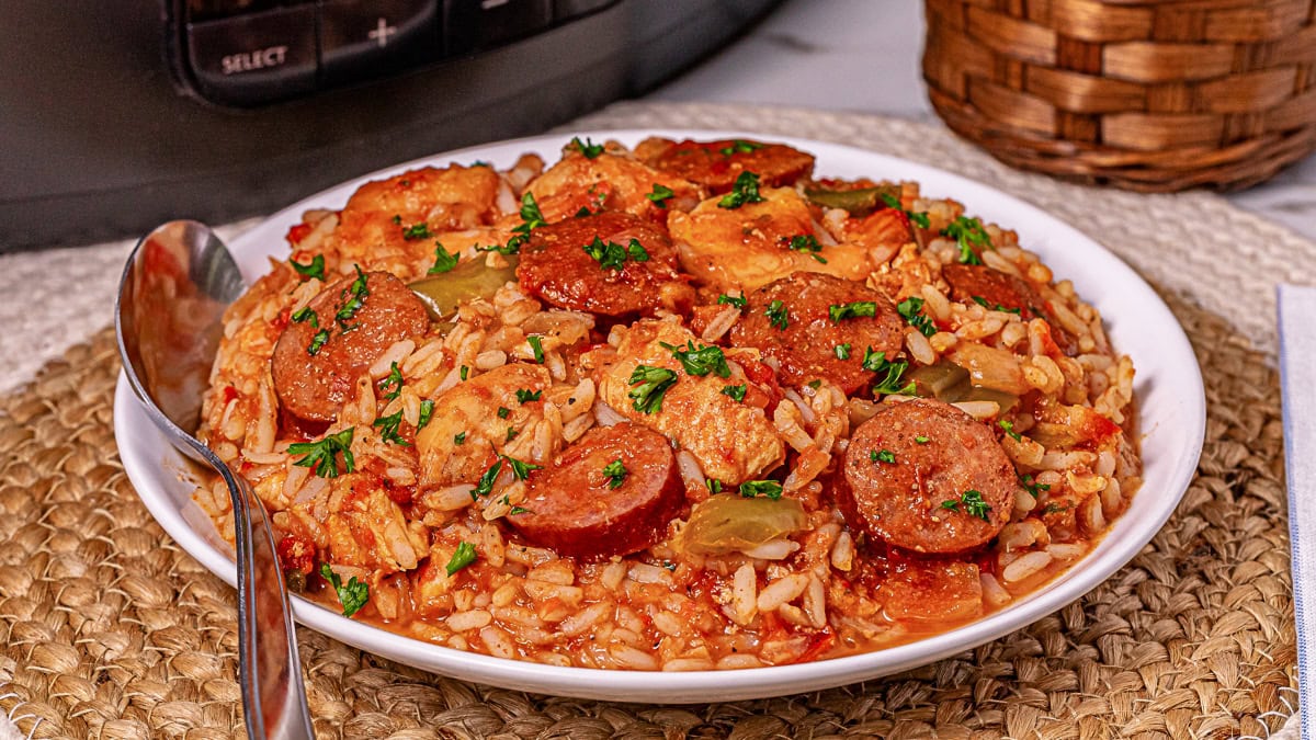 A white bowl filled with jambalaya containing rice, sausage, chicken, bell peppers, and tomato sauce, garnished with chopped parsley.
