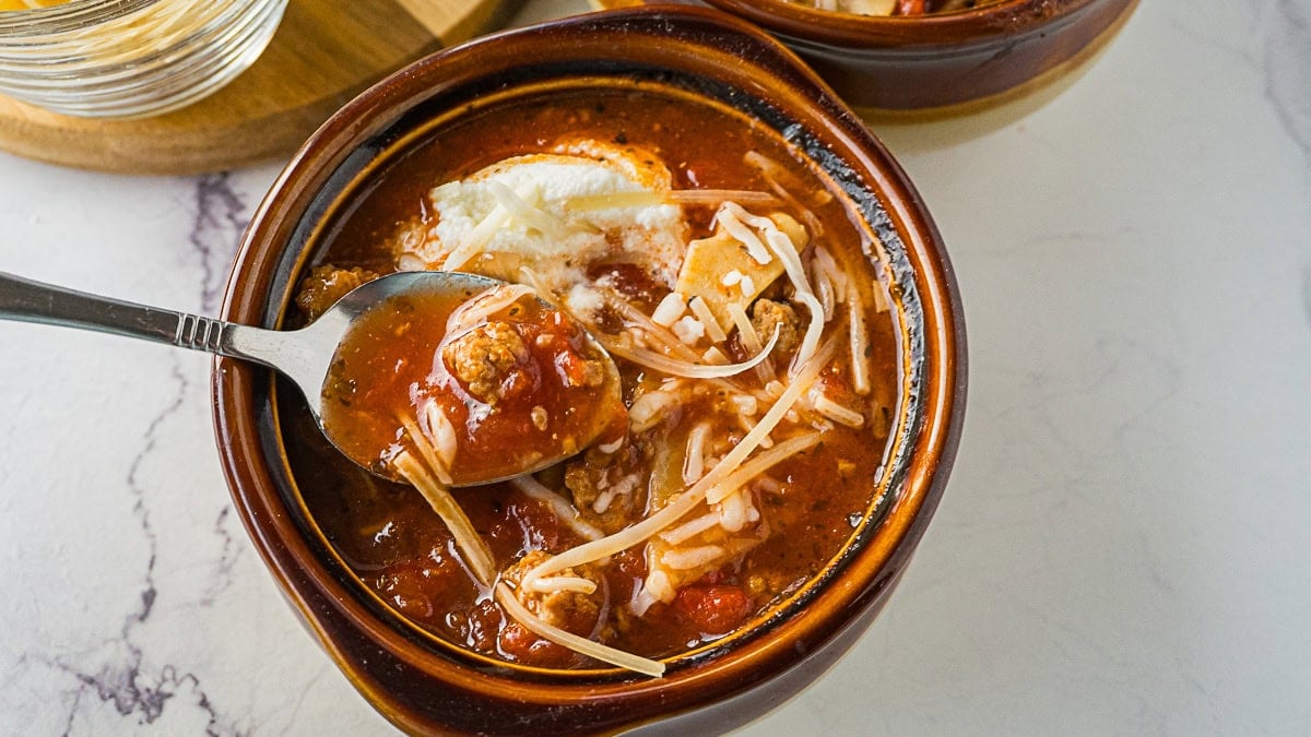 A brown bowl of lasagna soup topped with shredded cheese, with a spoon lifting a portion of soup.