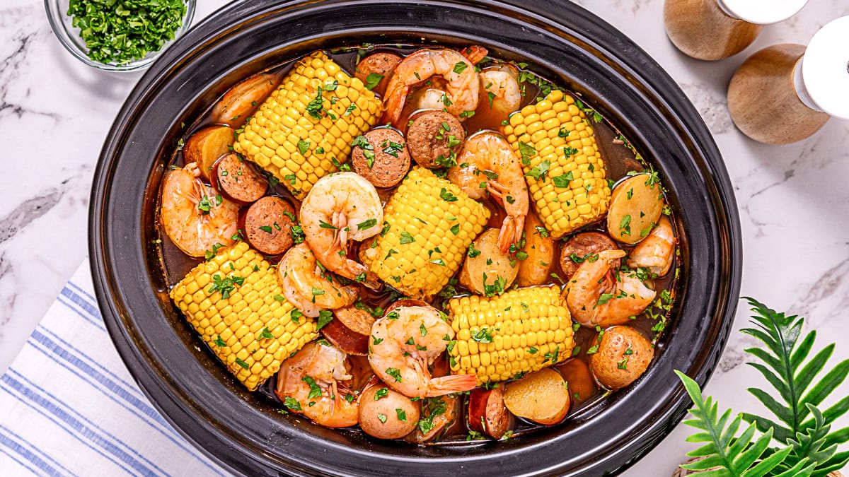 A slow cooker filled with shrimp, sausage slices, corn on the cob, and potatoes in broth, topped with chopped herbs.
