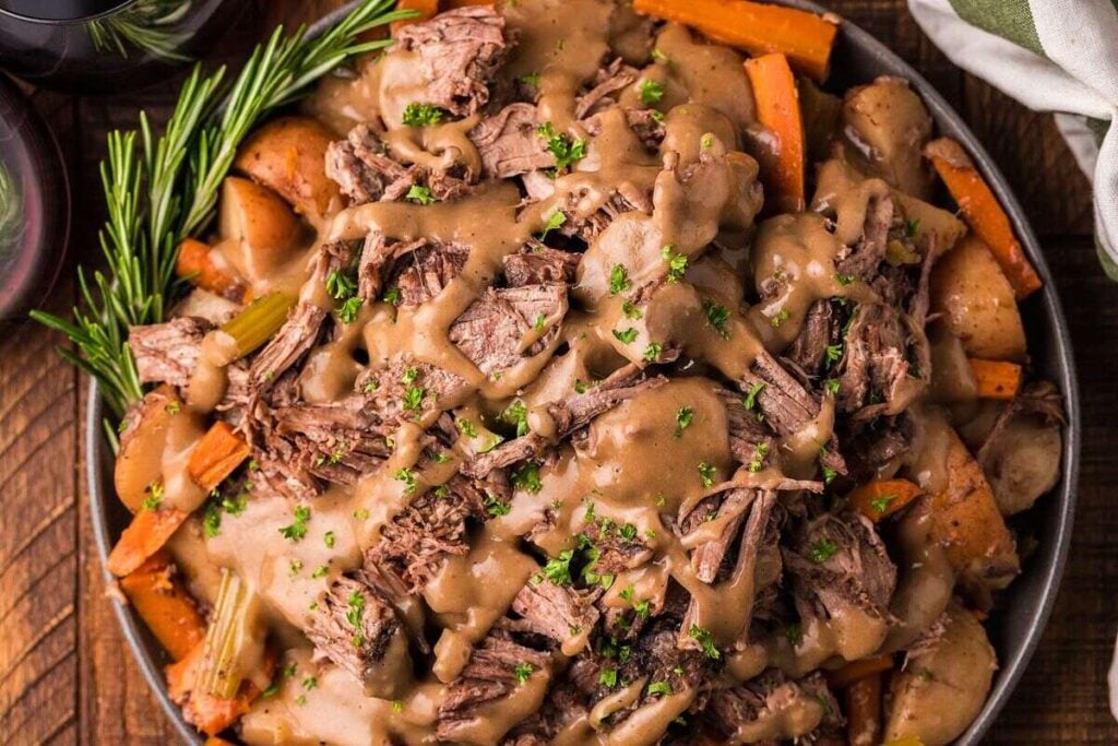 A bowl of pot roast with gravy, garnished with chopped parsley, served over carrots, potatoes, and celery, with a sprig of rosemary on the side.