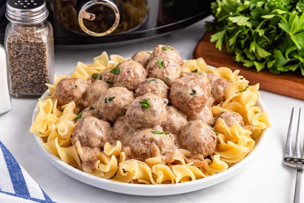 A plate of egg noodles topped with creamy sauce-covered meatballs, garnished with chopped parsley, sits on a white table near black pepper, fresh herbs, and a fork.