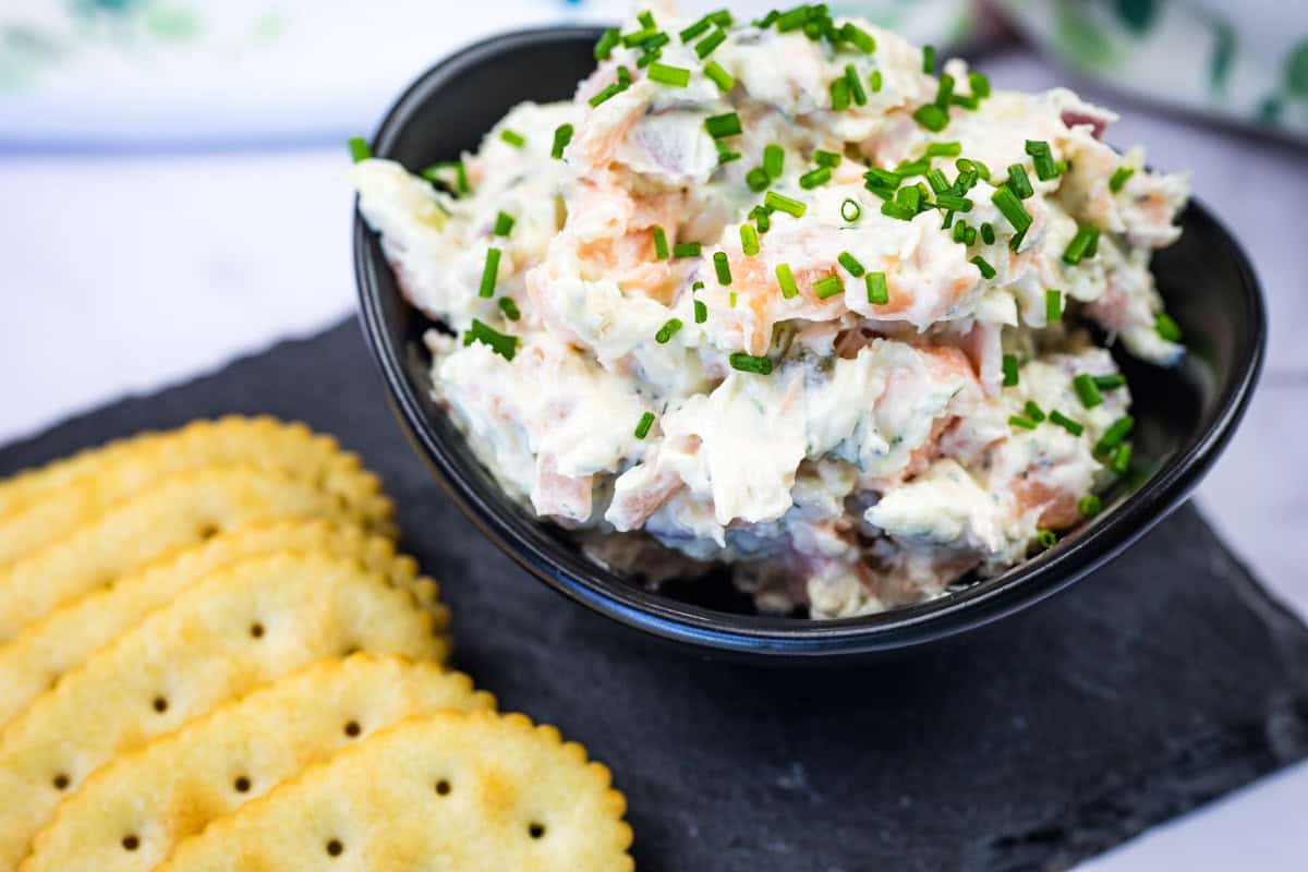Bowl of smoked salmon spread garnished with chopped chives next to crackers on a slate platter.