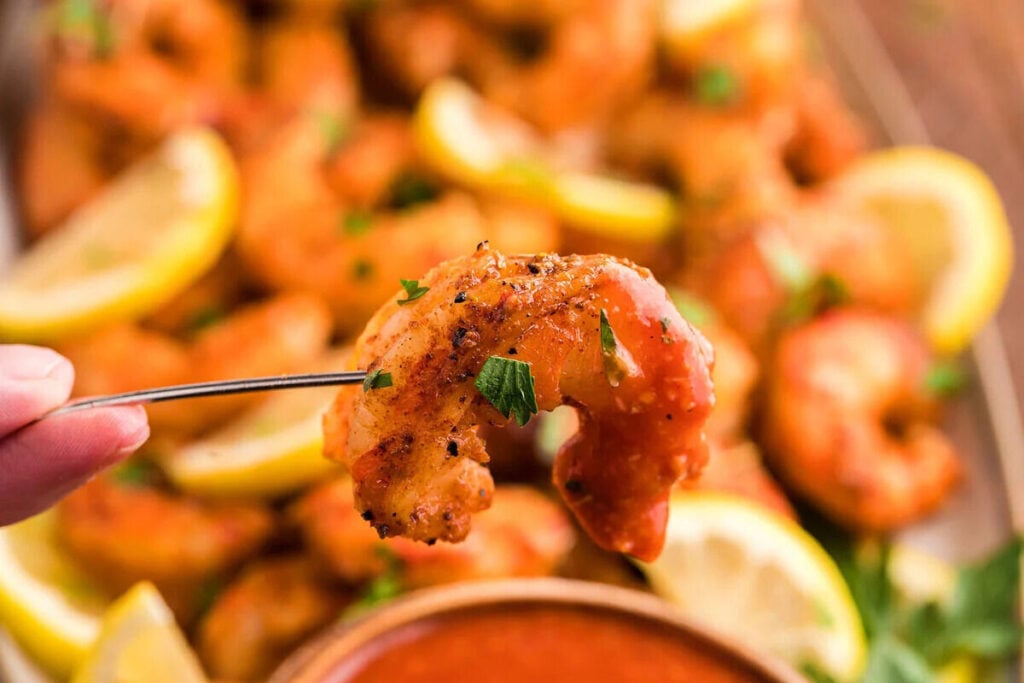 A hand holds a grilled shrimp on a fork above a platter of tasty appetizers featuring shrimp, lemon slices, and parsley, with a dish of red dipping sauce nearby.
