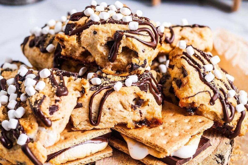 A pile of s’mores-inspired dessert bars topped with mini marshmallows and chocolate drizzle, stacked on top of classic s’mores with graham crackers, chocolate, and melted marshmallows.