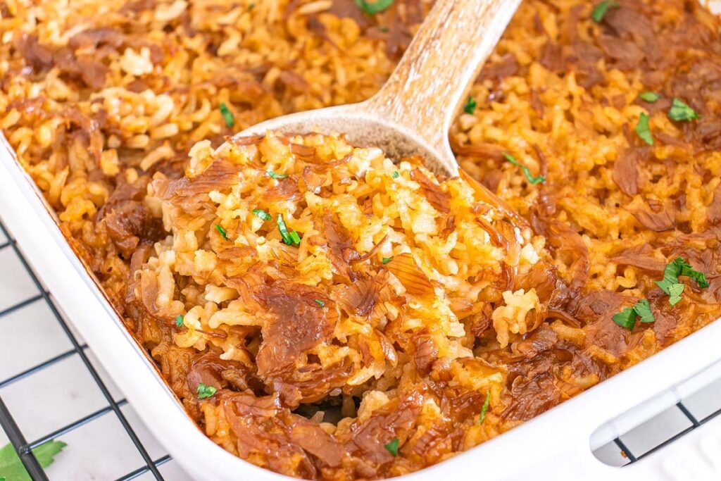 A close-up of a baked rice casserole with shredded meat, topped with herbs, in a white dish. A wooden spoon is lifting a portion.