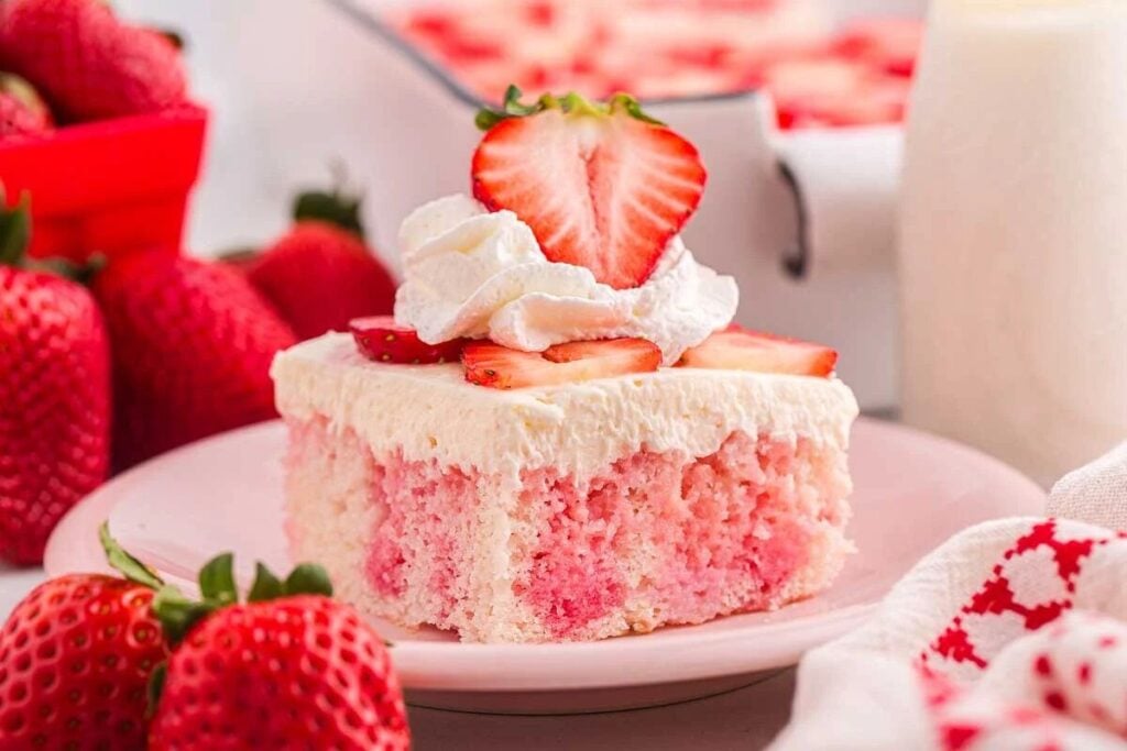 A slice of strawberry poke cake topped with whipped cream and sliced strawberries on a pink plate, surrounded by fresh strawberries.