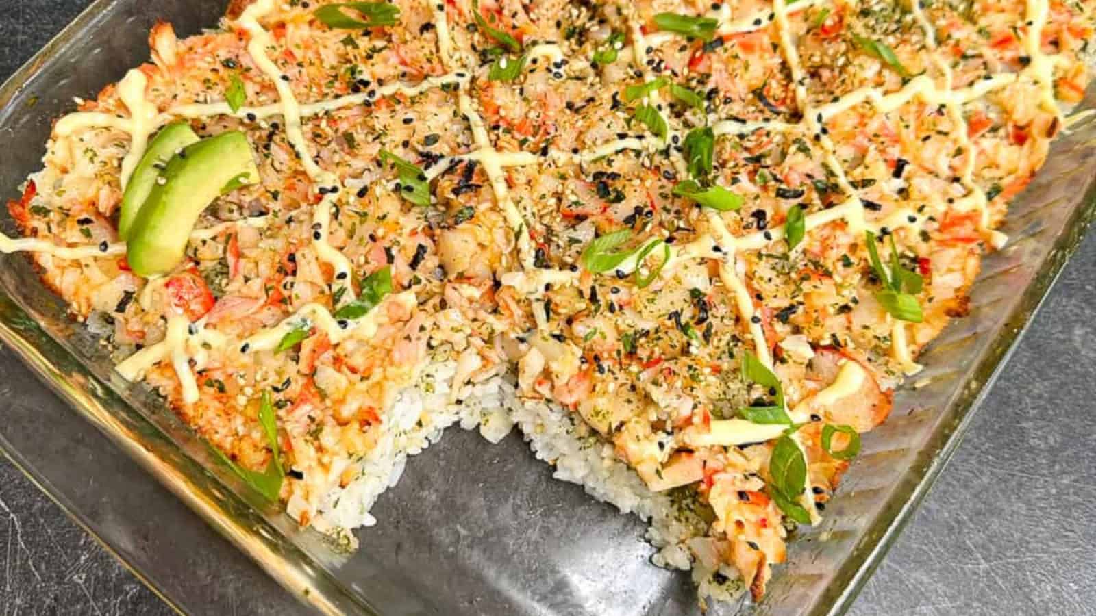 A baking dish filled with sushi bake, featuring layers of rice, seafood topping, mayonnaise drizzle, avocado slices, sesame seeds, and herbs.