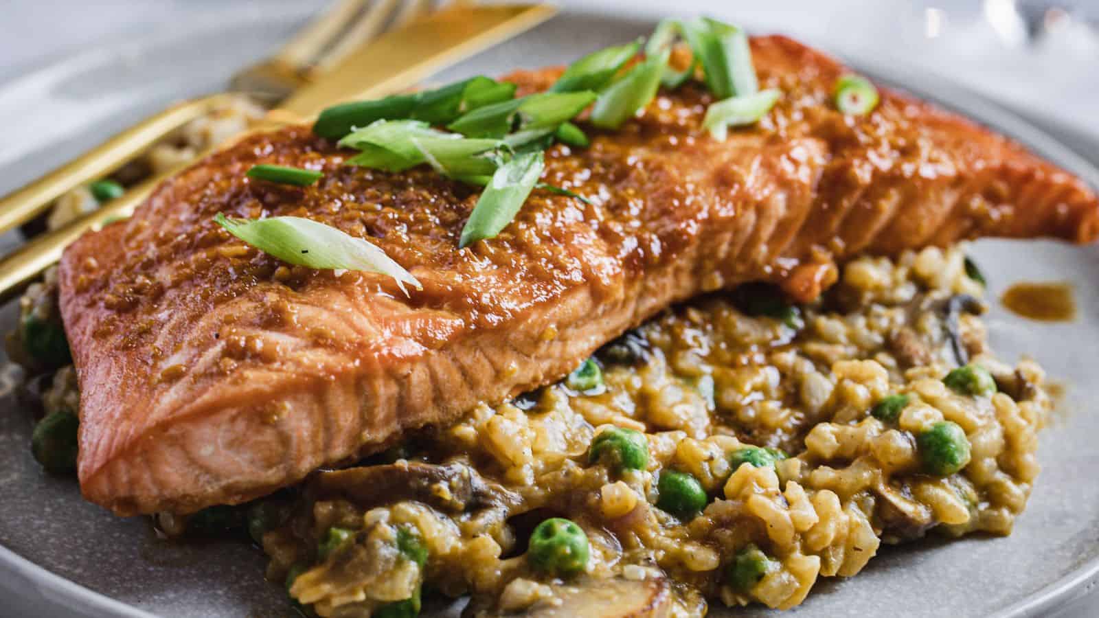 A cooked salmon fillet sits on top of a bed of risotto with peas and mushrooms, garnished with sliced green onions, on a round plate with a fork and knife.