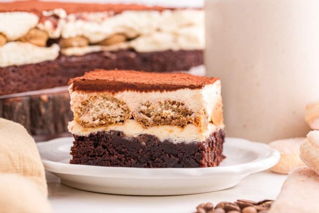A close-up of a slice of layered tiramisu dessert on a white plate, showing distinct layers of cream, cake, and cocoa powder.