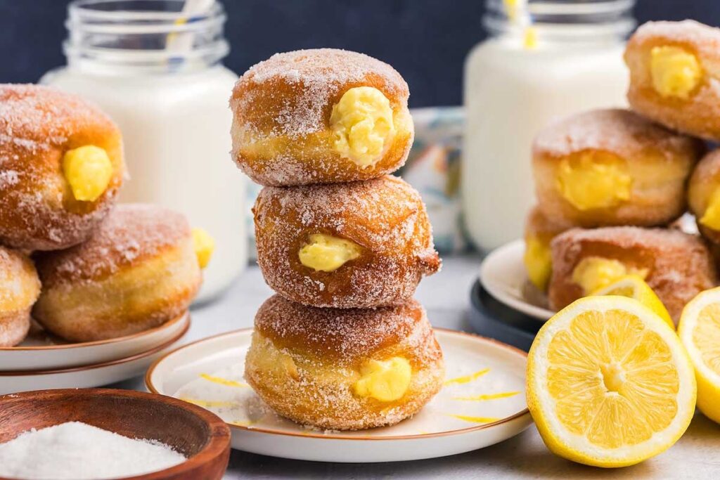 A stack of three sugar-coated bomboloni filled with lemon custard, surrounded by more custard-filled doughnuts, a halved lemon, a bowl of sugar, and jars of milk with straws in the background.