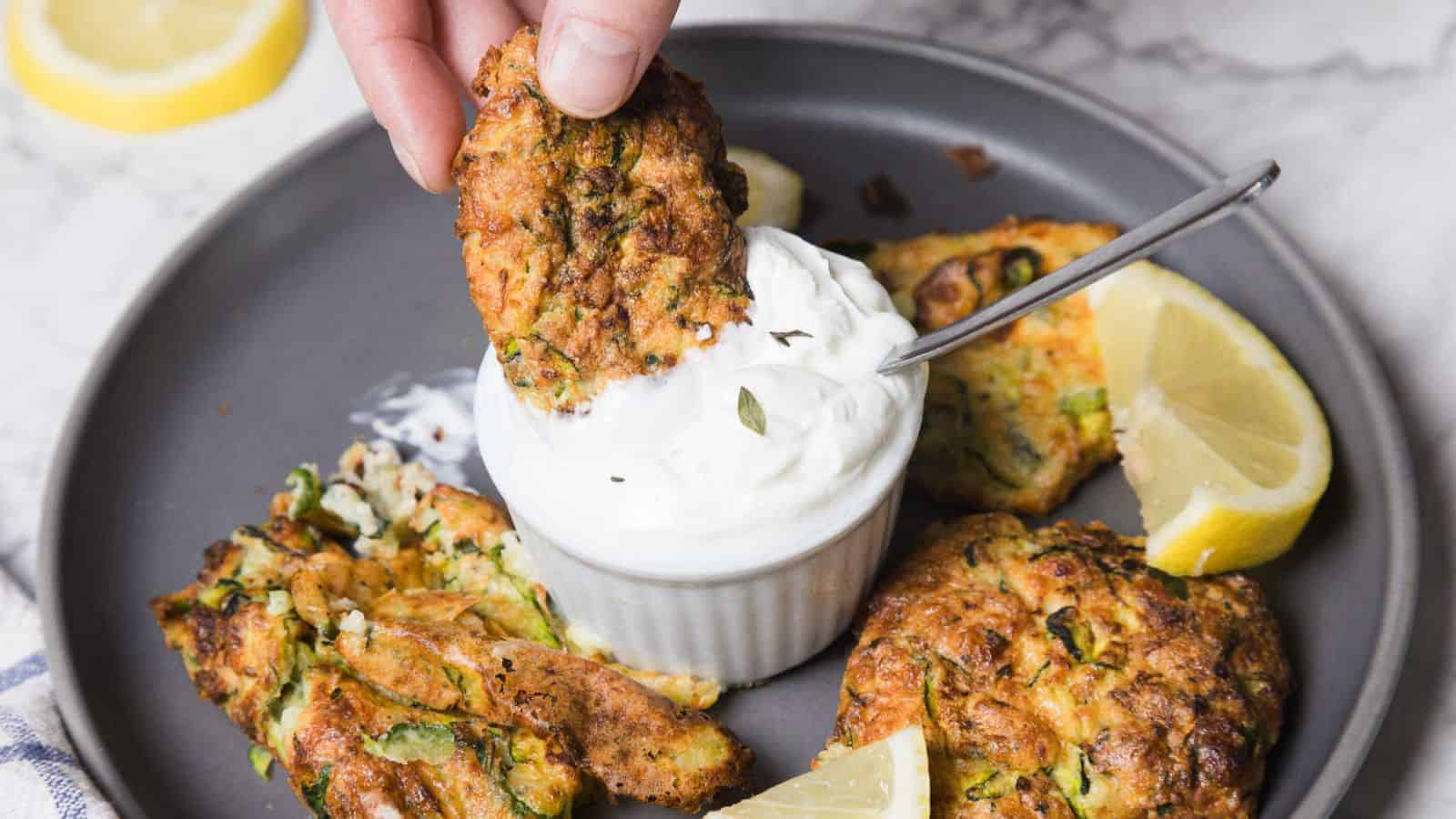 A hand dips a fried zucchini fritter into a small bowl of white cream sauce on a gray plate. Other fritters and lemon wedges are arranged around the bowl on a marble surface.