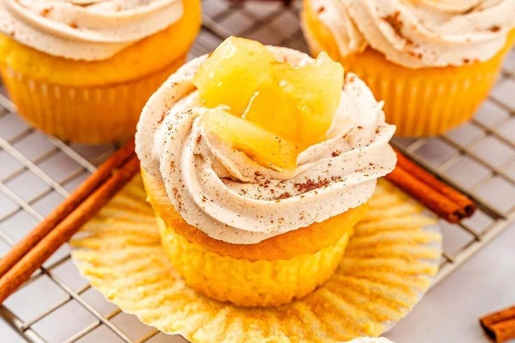 A yellow cupcake with swirled cream frosting topped with pineapple chunks, dusted with cinnamon, sits on an open wrapper, surrounded by cinnamon sticks.