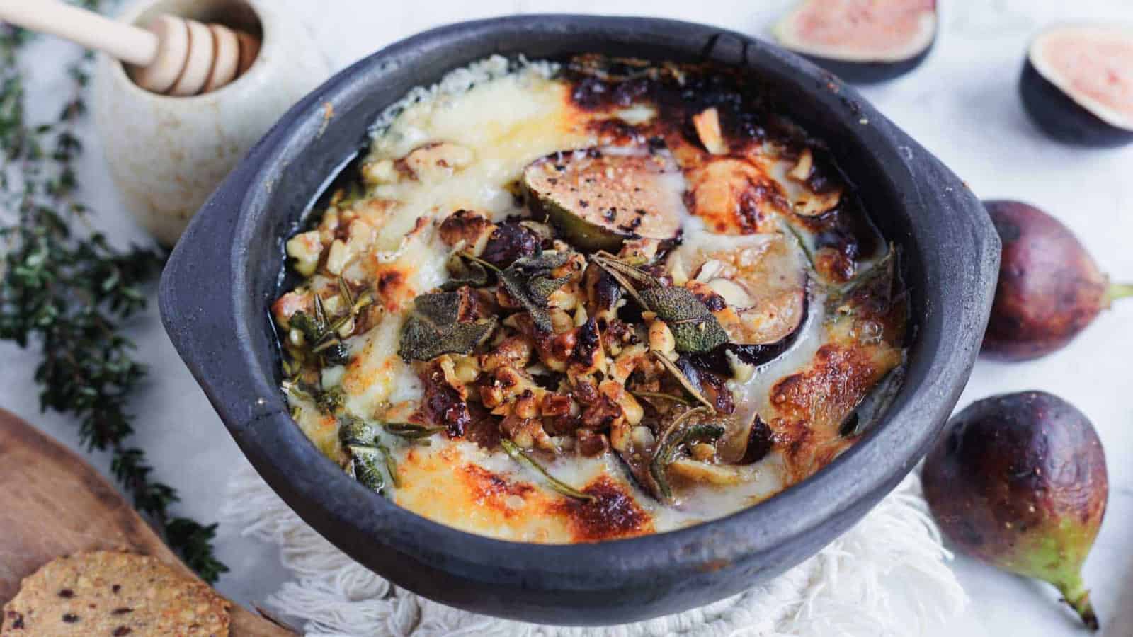Baked cheese dish with figs, herbs, and nuts in a black bowl.
