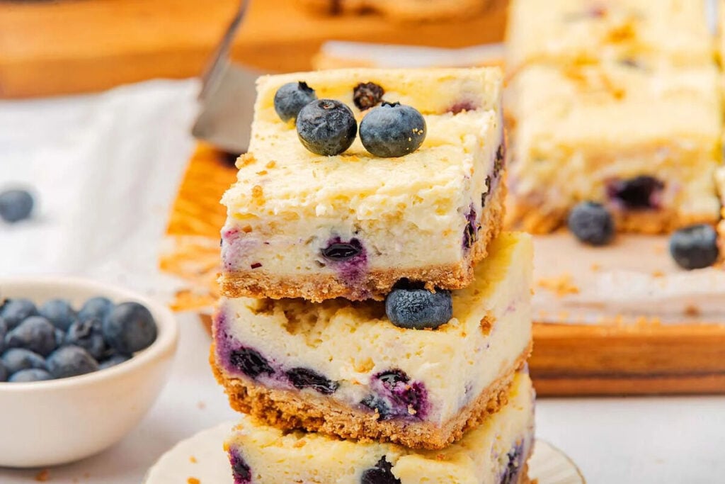 Three stacked blueberry cheesecake bars with a crumbly crust, topped with fresh blueberries. 