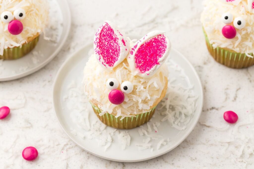 A bunny cupcake with pink ears, candy eyes, and a pink nose sits on a white plate.