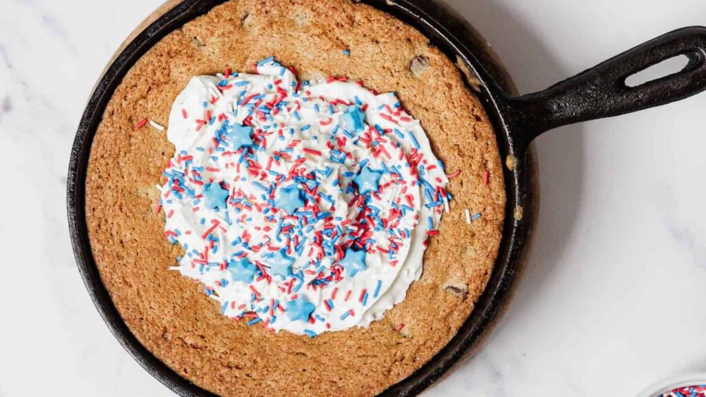 A large chocolate chip cookie baked in a cast iron skillet, topped with white frosting and red, white, and blue sprinkles, viewed from above on a white surface.