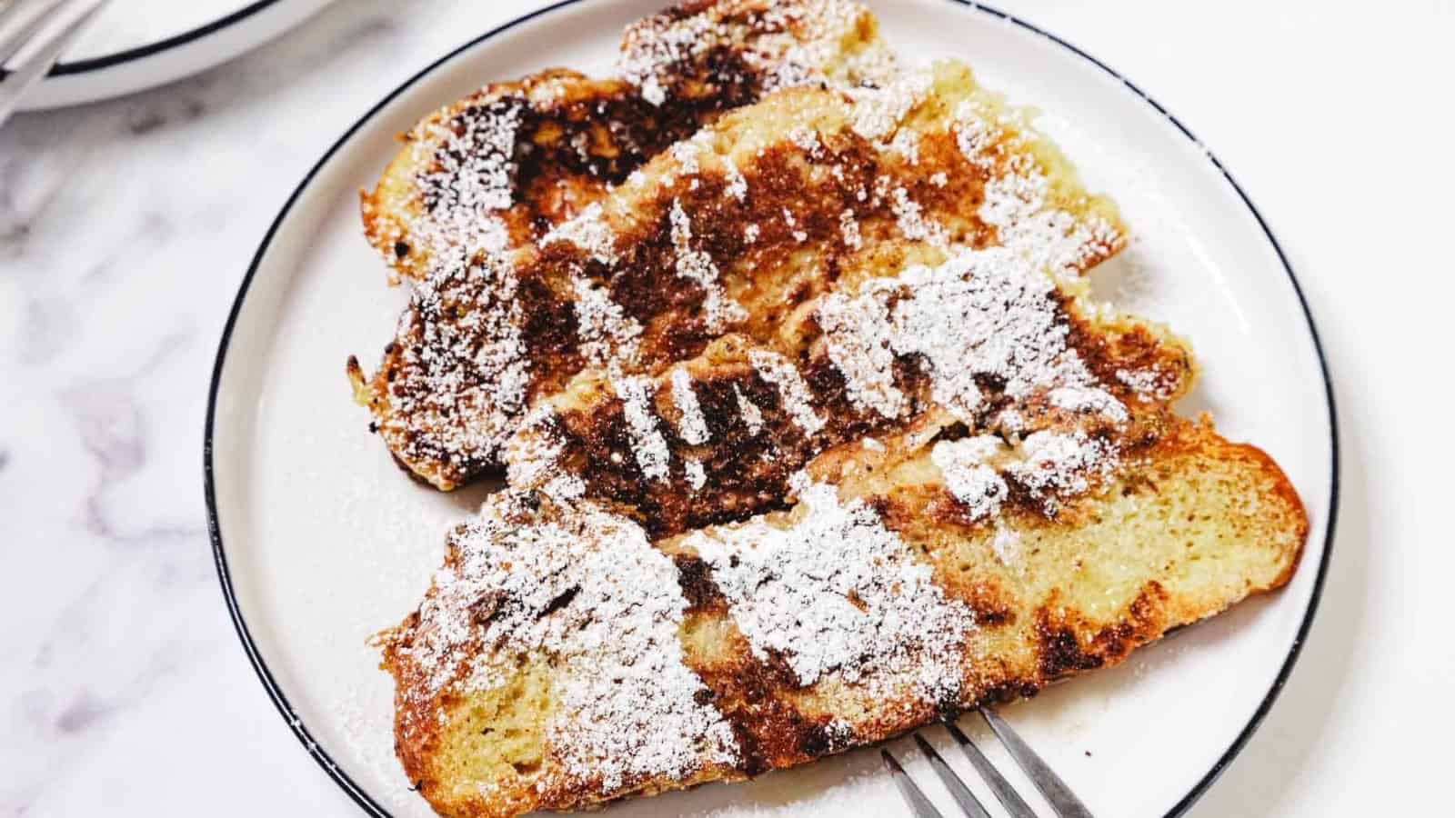 Four slices of French toast topped with powdered sugar on a white plate, with a fork beside them.