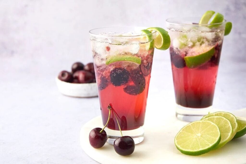 Two glasses of Cherry Caipirinha, a vibrant Brazilian cocktail made with Cachaça or dark rum, ice, and garnished with lime slices, cherries, and fresh lime nearby.