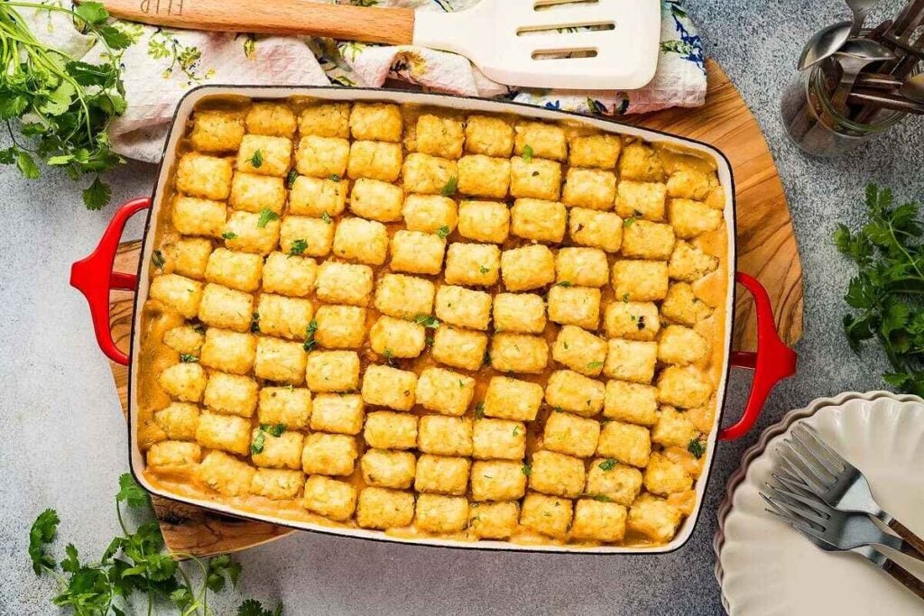 A baking dish filled with neatly arranged tater tots on top of a creamy chicken tater tot casserole, placed on a wooden board with utensils and cilantro garnish nearby.