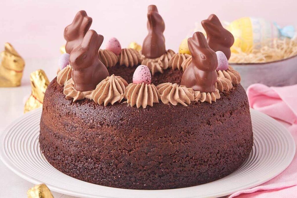 Chocolate Easter Bundt Cake topped with chocolate bunnies and mini eggs sits elegantly on a white plate, surrounded by festive Easter decor.