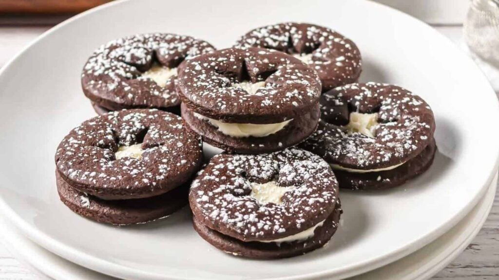 Chocolate sandwich cookies with cream filling, dusted with powdered sugar, arranged on a white plate.
