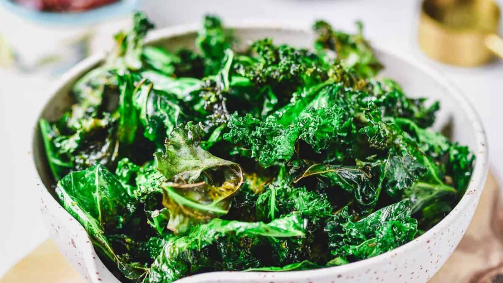 A white speckled bowl filled with crispy, dark green kale chips sits on a light surface. The kale chips appear roasted with some edges curled and slightly browned.