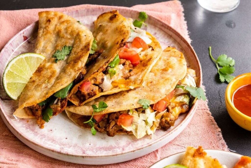  Three crispy turkey tacos filled with meat, lettuce, tomato, and cilantro, served with lime on a plate.
