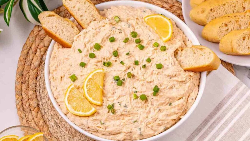A bowl of creamy dip garnished with lemon slices and green onions, served with slices of baguette.