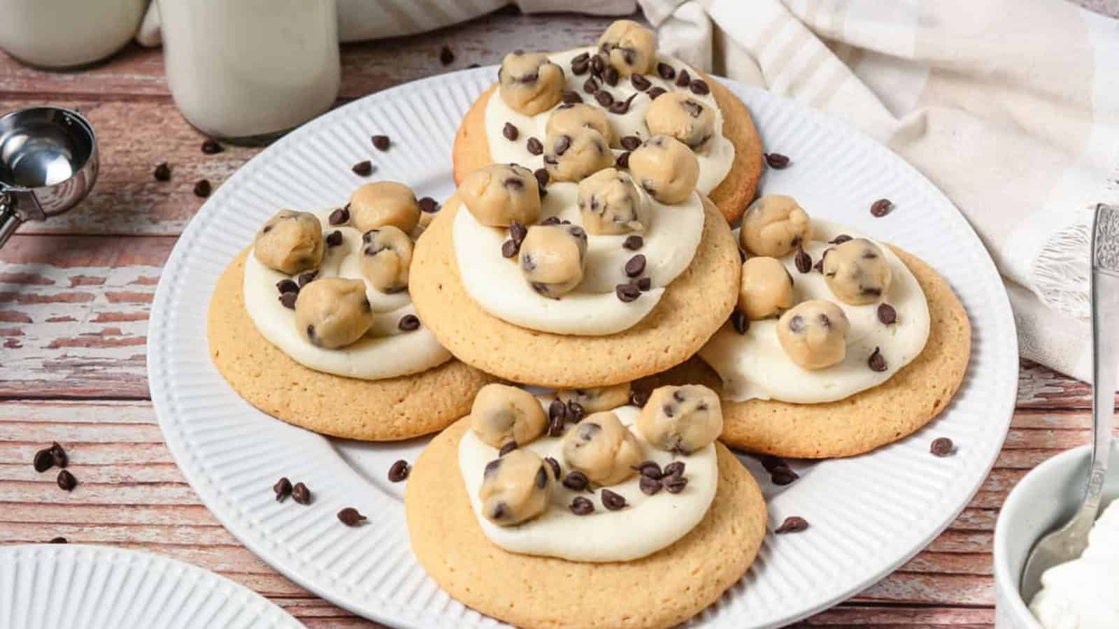 Five cookies topped with frosting, mini chocolate chip cookie dough balls, and chocolate chips on a white plate.