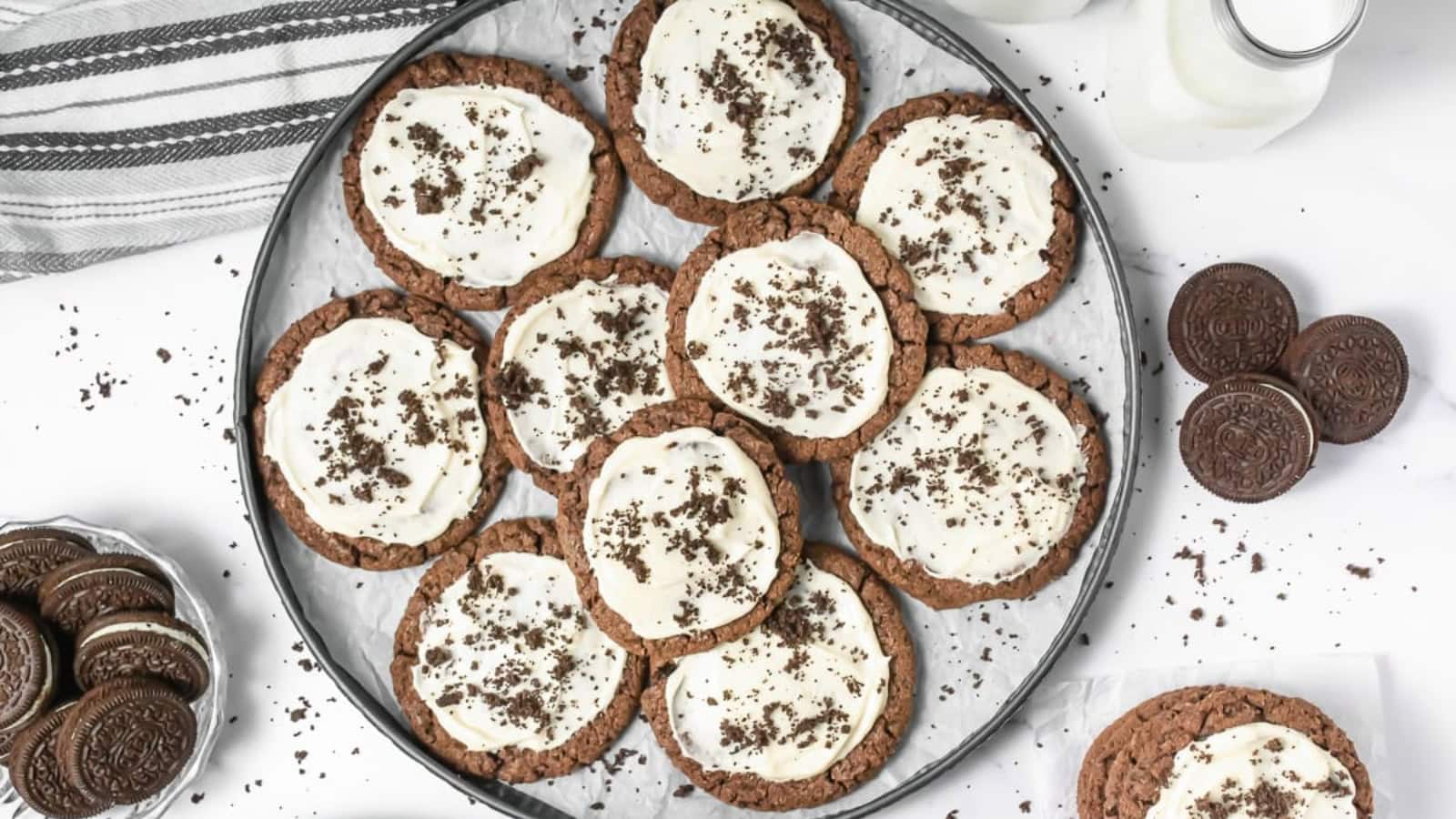 A plate of chocolate cookies with white icing and cookie crumbs, surrounded by sandwich cookies and milk.