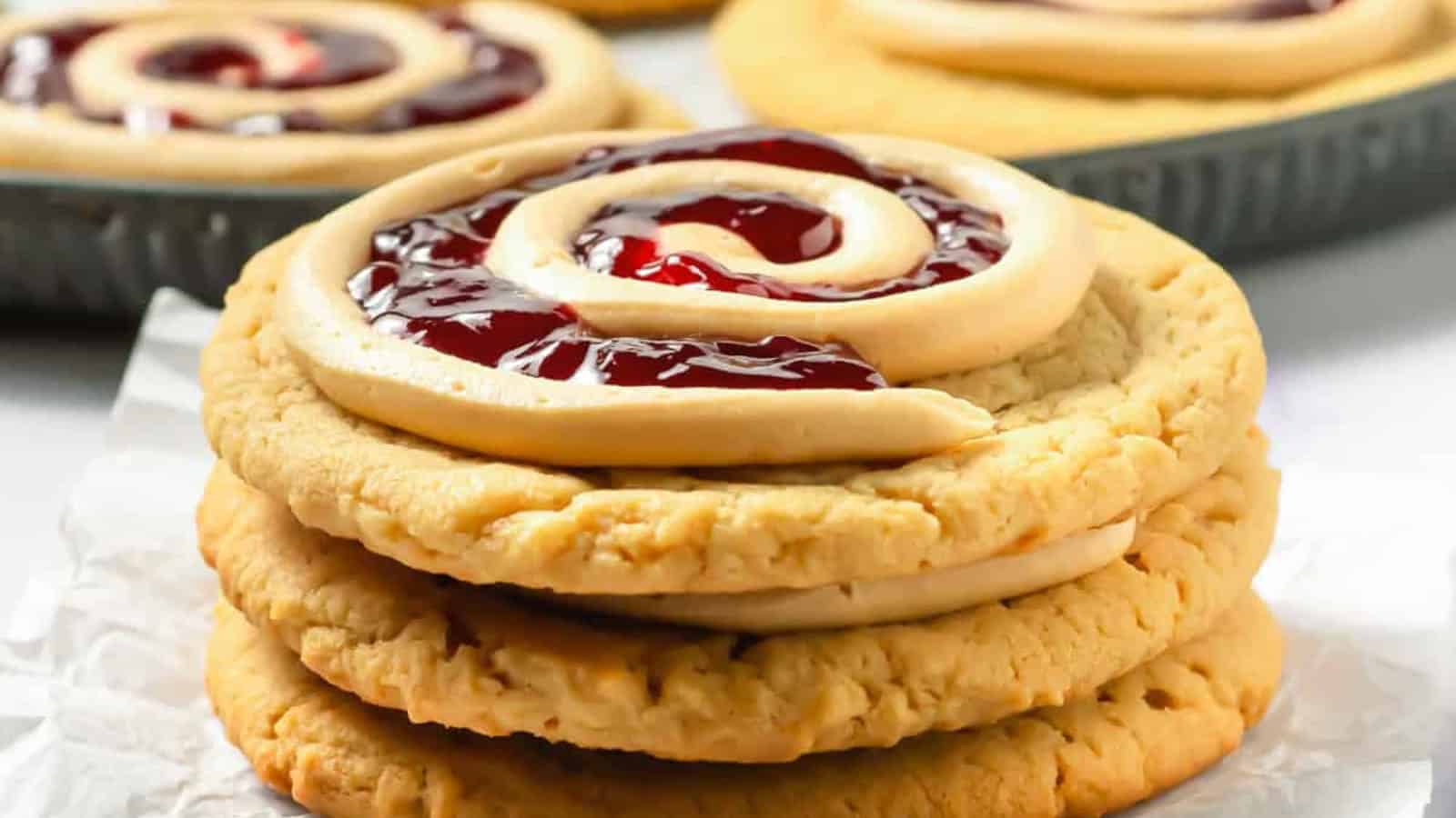 A stack of cookies topped with a swirl of icing and red jelly filling, with more cookies in the background.