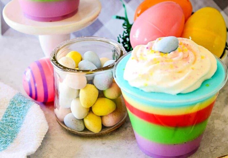 A close-up of a layered rainbow-colored dessert in a plastic cup, topped with a swirl of whipped cream and a small blue candy. To the left is a glass jar filled with pastel-colored egg-shaped candies, and behind are plastic Easter eggs.