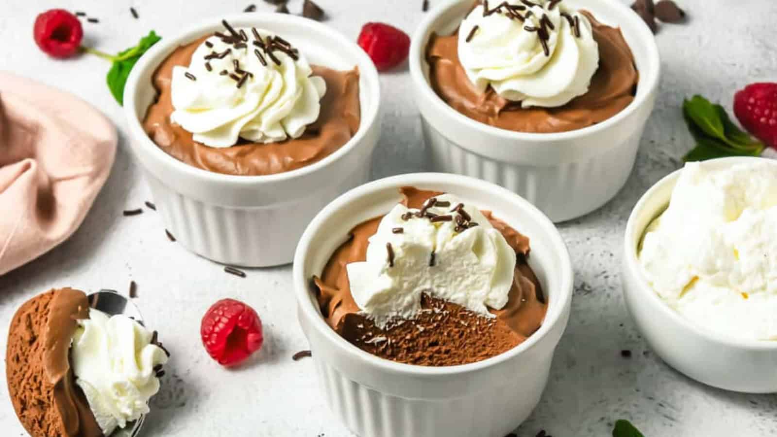 Four ramekins with chocolate mousse topped with whipped cream and chocolate shavings, surrounded by raspberries.