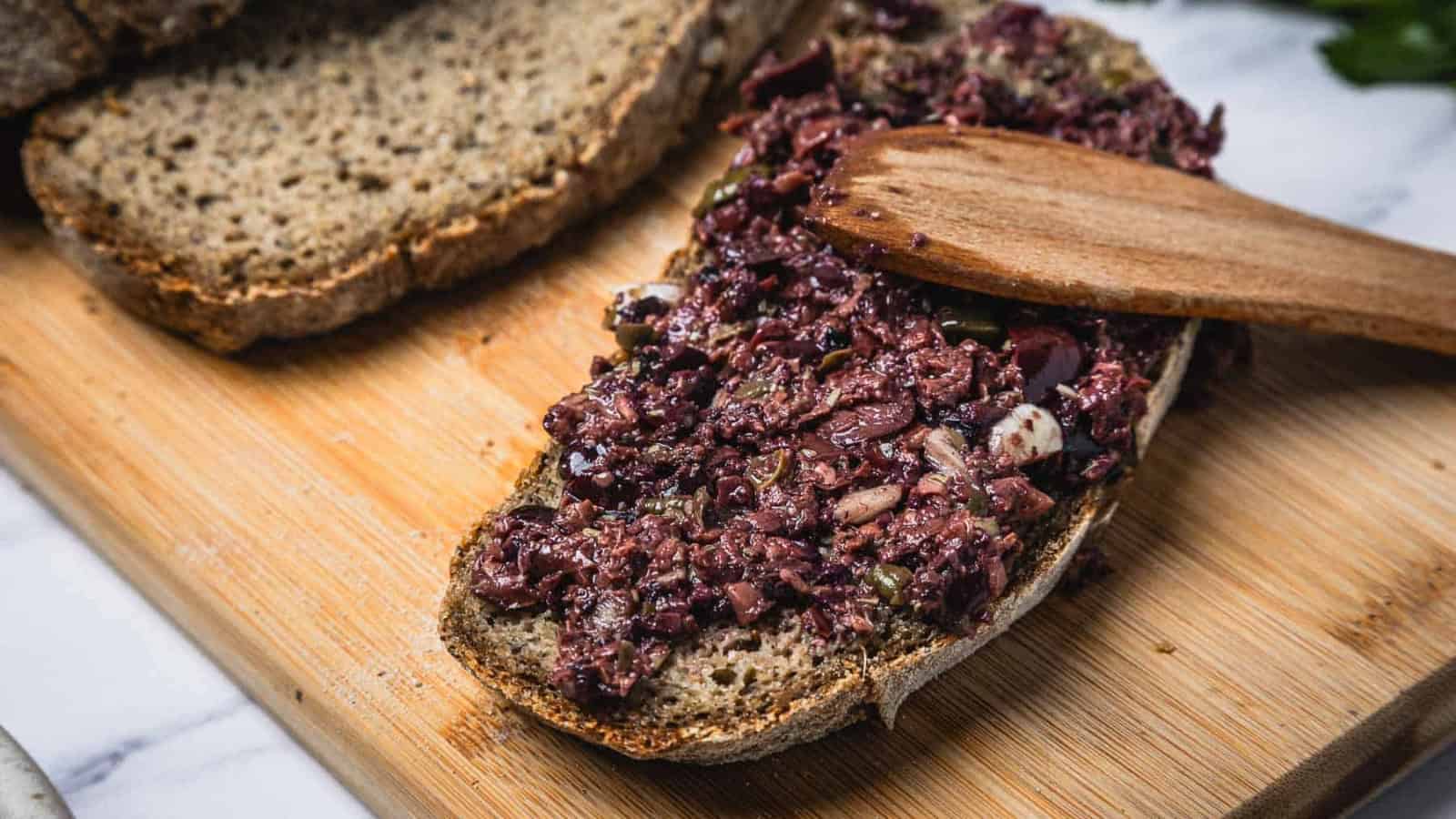 A slice of bread topped with olive tapenade being spread with a wooden spatula on a cutting board.