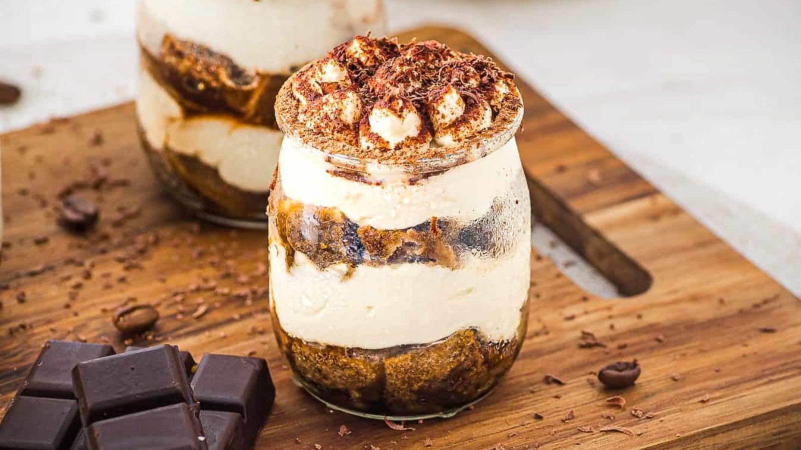 Two glass jars of tiramisu topped with cocoa and chocolate shavings, next to pieces of dark chocolate.