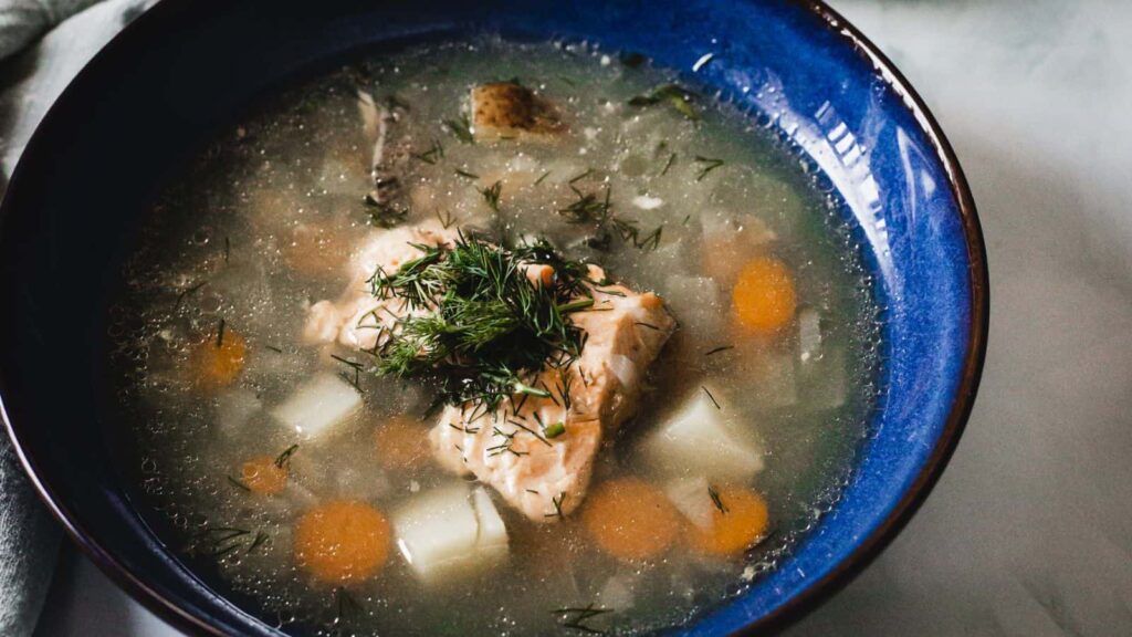A blue bowl contains a soup with chunks of fish, carrots, and potatoes, garnished with fresh dill. The soup appears to have a clear broth base.