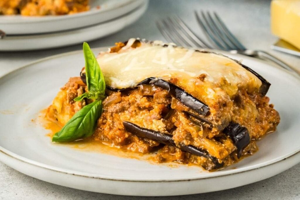 A serving of eggplant lasagna topped with melted cheese and garnished with a basil leaf on a white plate with a fork in the background.