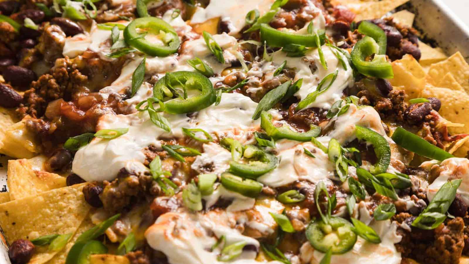 A close-up of nachos topped with melted cheese, ground beef, sliced jalapeños, chopped green onions, sour cream, and beans. The tortilla chips are spread out with the toppings generously applied, creating a colorful and appetizing presentation.